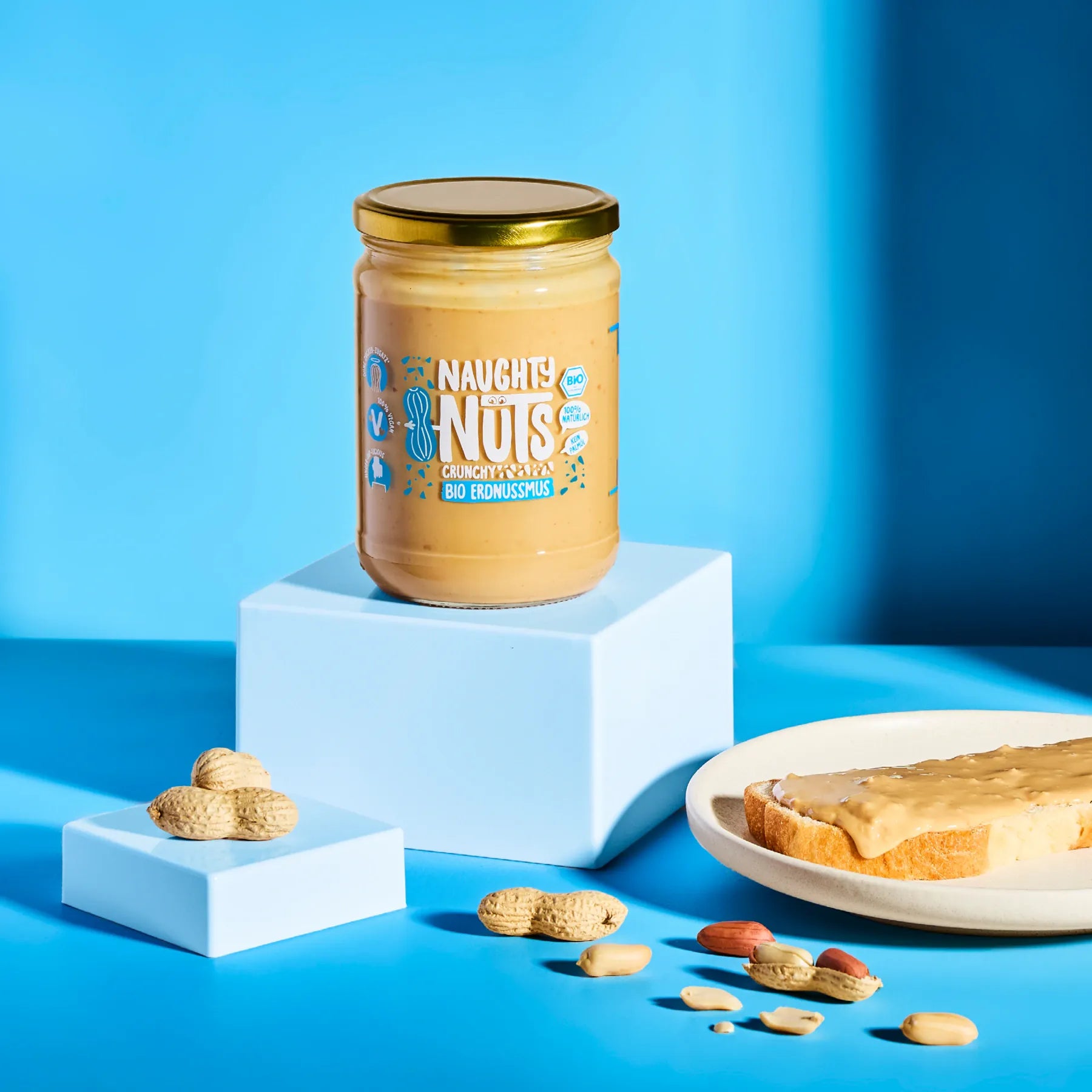 Bio Erdnussmus Crunchy im Glas auf einem Podest mit Erdnüssen und einer Scheibe Brot