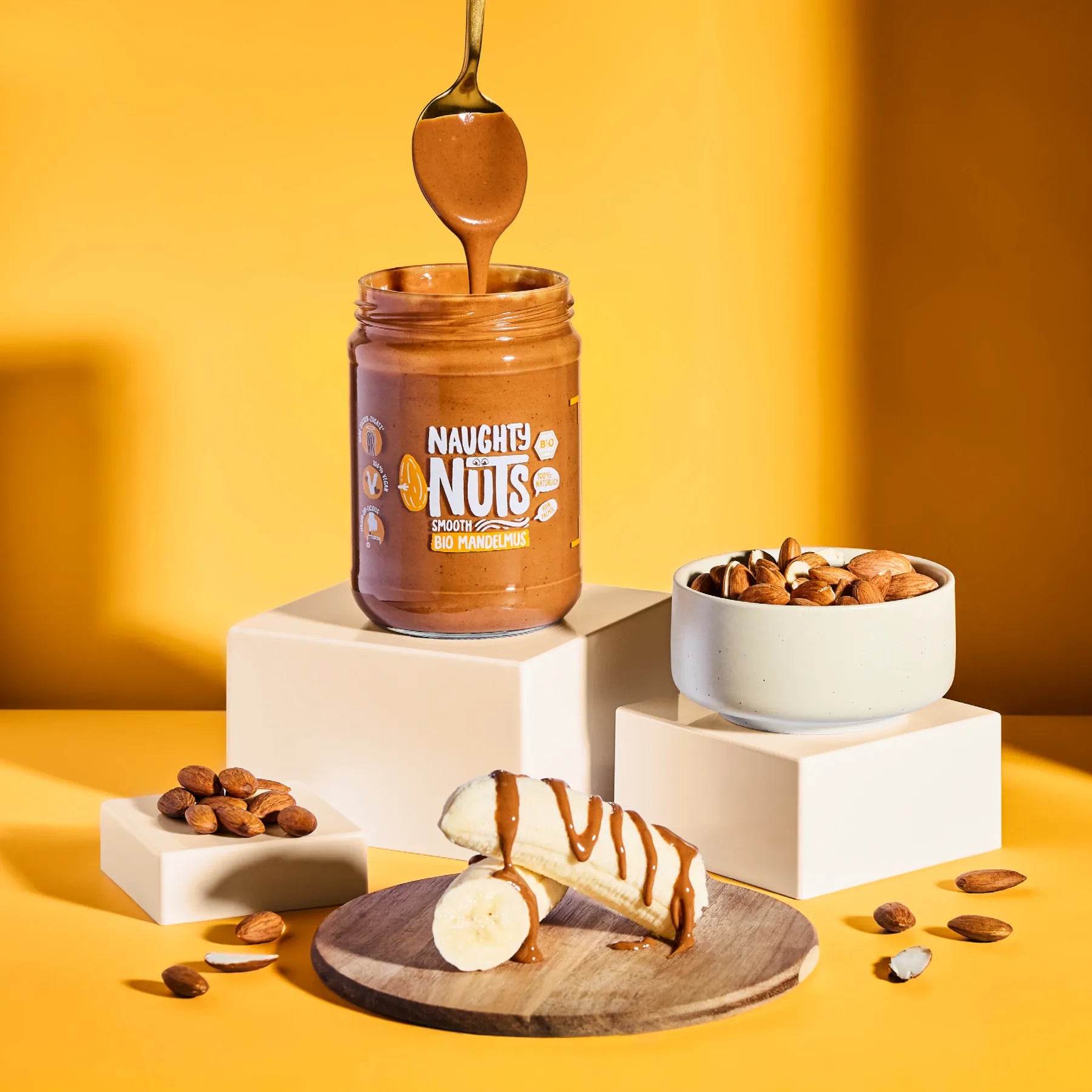 Bio Mandelmus Smooth im Glas mit Löffel, Bananenstücken und Mandeln auf gelbem Hintergrund